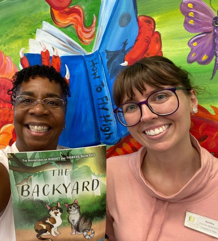 Two women standing in front of a mural with a book in front of them.