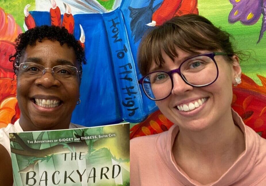 Two women standing in front of a mural with a book in front of them.